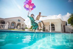 Happy young couple jumping into the pool while holding a bunch of balloons